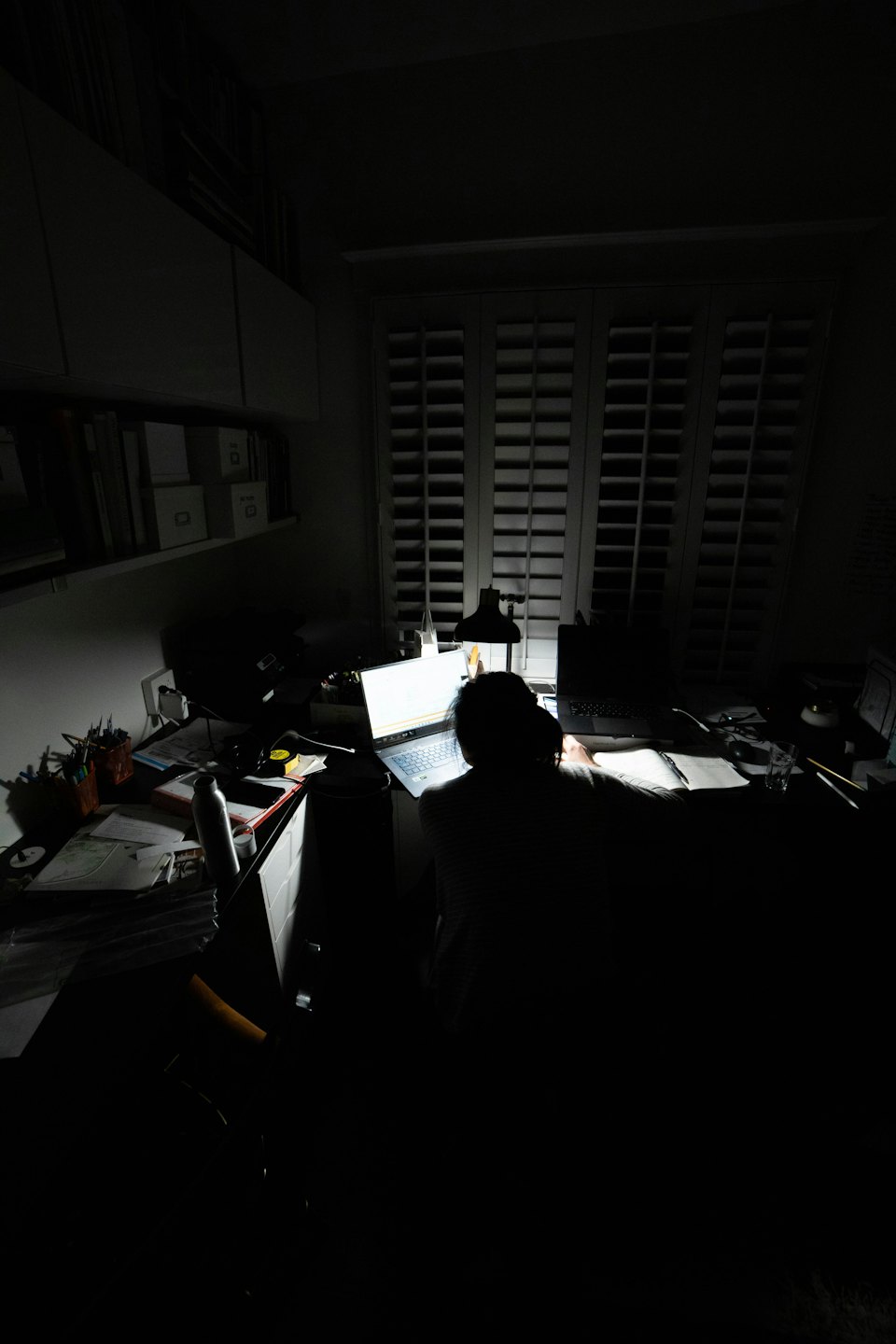 Person working on laptop at a cluttered desk in a dark room.